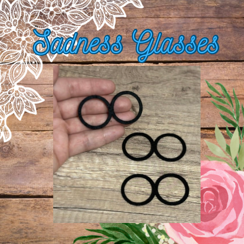 A pair of black felt glasses held in a hand, with the text 'SADNESS GLASSES' and a floral pattern in the background.