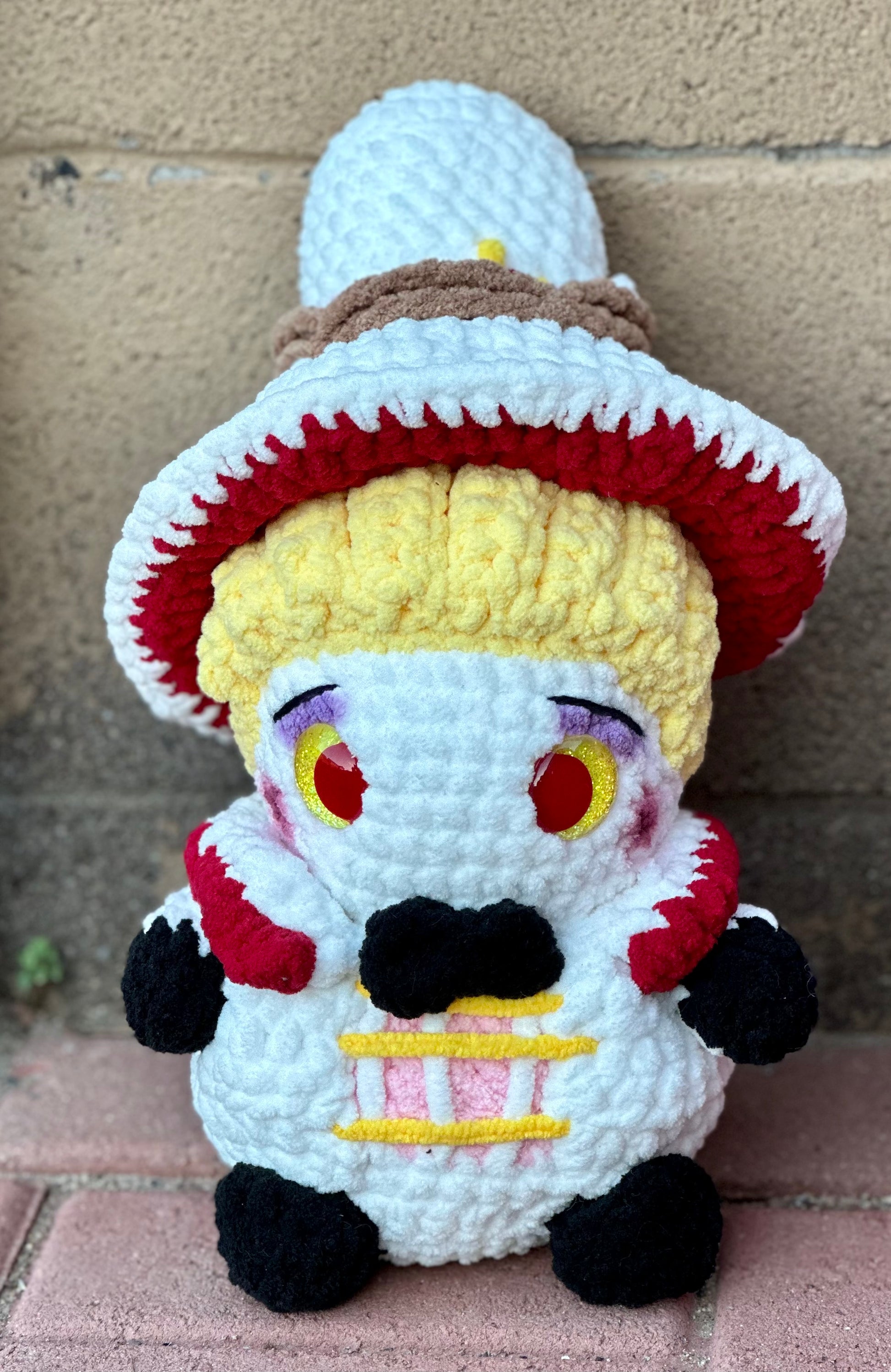 A crochet plush character with a large white and red hat, yellow hair, and red and yellow eyes sits outdoors against a brick wall. The plush is handmade with chunky yarn in white, red, black, and yellow, featuring small arms, black hands and feet, and a bow tie